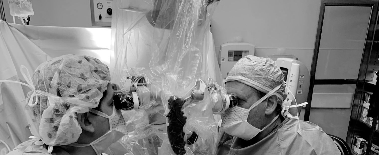 Two surgeons working through an operating microscope, heads lowered to the eyepieces.