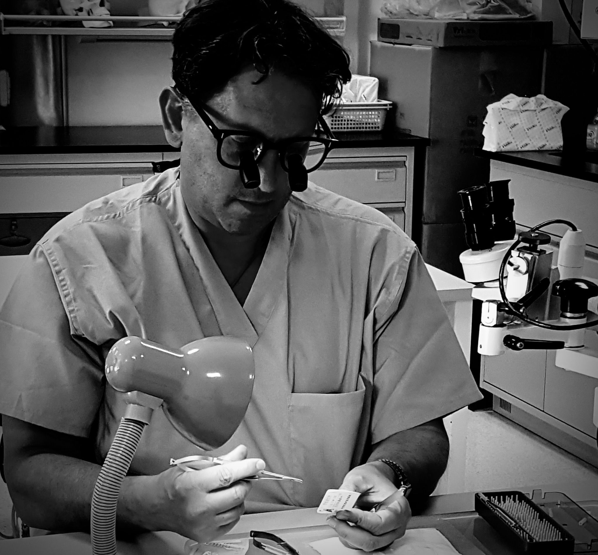 Andrew Mark Klapper at a microsurgical laboratory bench, wearing loupes and holding microsurgical instruments.