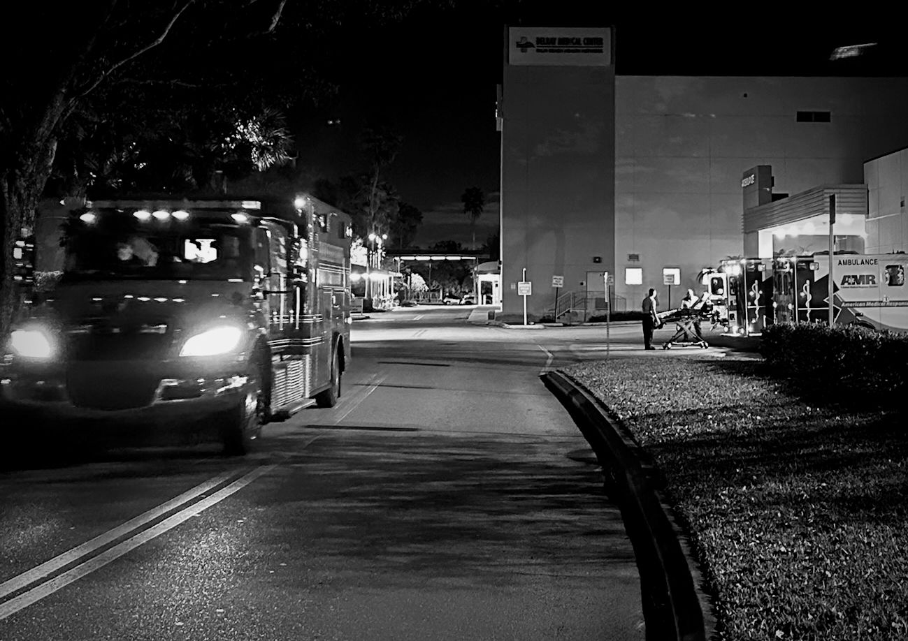 Night view of a hospital emergency entrance: a fire rescue engine approaching along the roadway at left, and an ambulance with a stretcher team at the bay on the right.